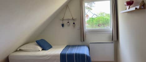 Un dormitorio pequeño y acogedor con techo inclinado, una cama individual con colcha azul a rayas, una mesita y una ventana con vistas a zonas verdes.