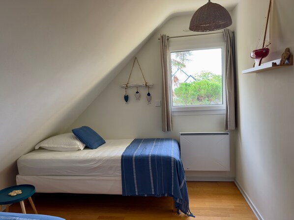 Un dormitorio pequeño y acogedor con techo inclinado, una cama individual con colcha azul a rayas, una mesita y una ventana con vistas a zonas verdes.