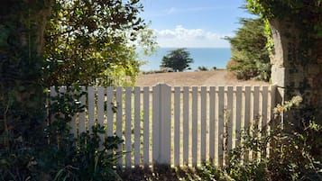 Ein rundbogiges Steintor mit weißem Zaun, das zu einem Strand mit freiem Blick auf das Meer und Bäume in der Ferne führt.