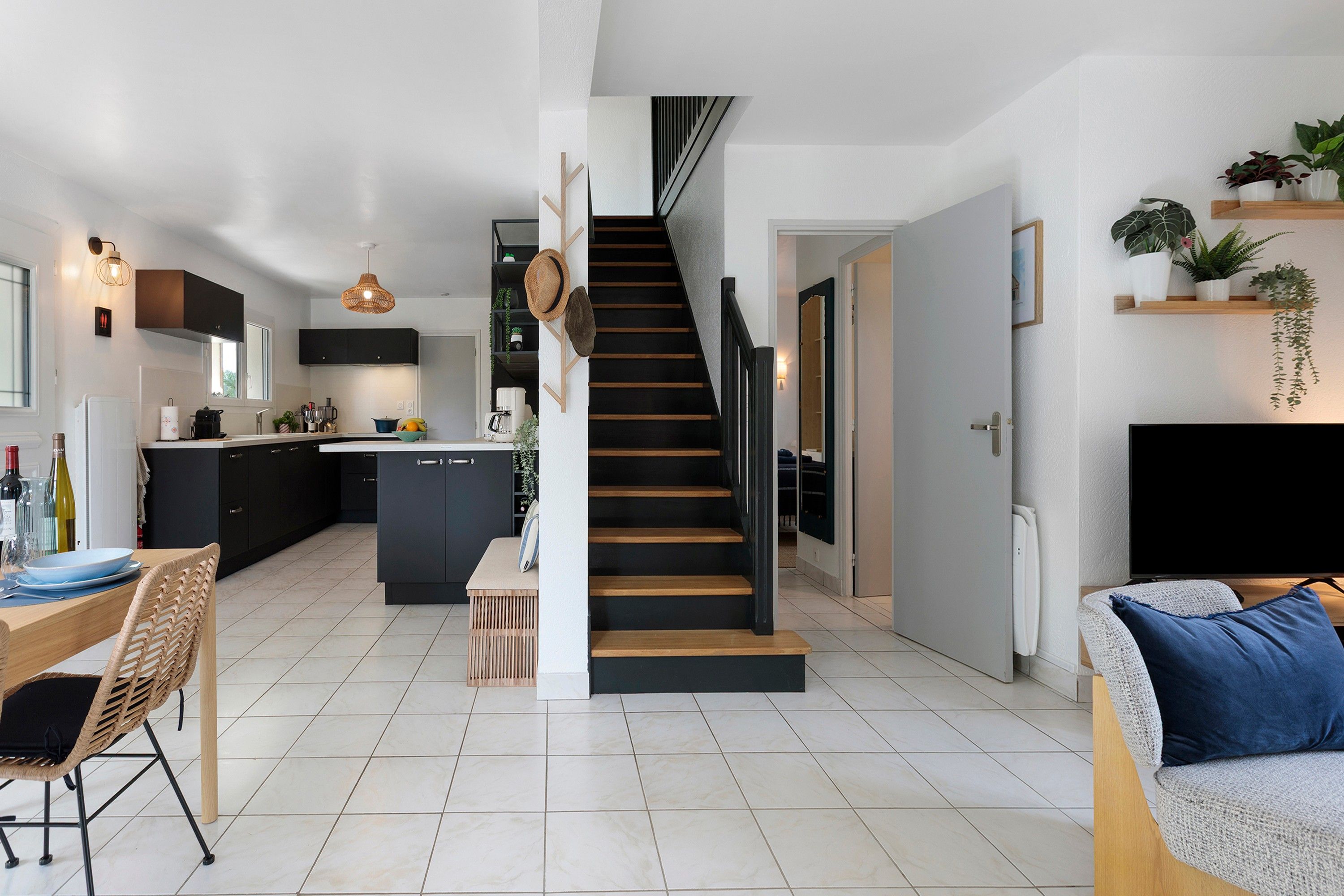 Une cuisine moderne avec des meubles de couleur sombre, un coin repas avec une table en bois et des chaises en osier, et un escalier menant à un autre niveau.