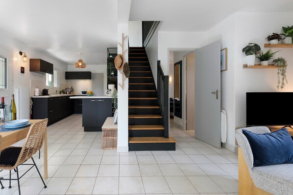 Una cocina moderna con armarios oscuros, un comedor con mesa de madera y sillas de mimbre, y una escalera que conduce a otro nivel.