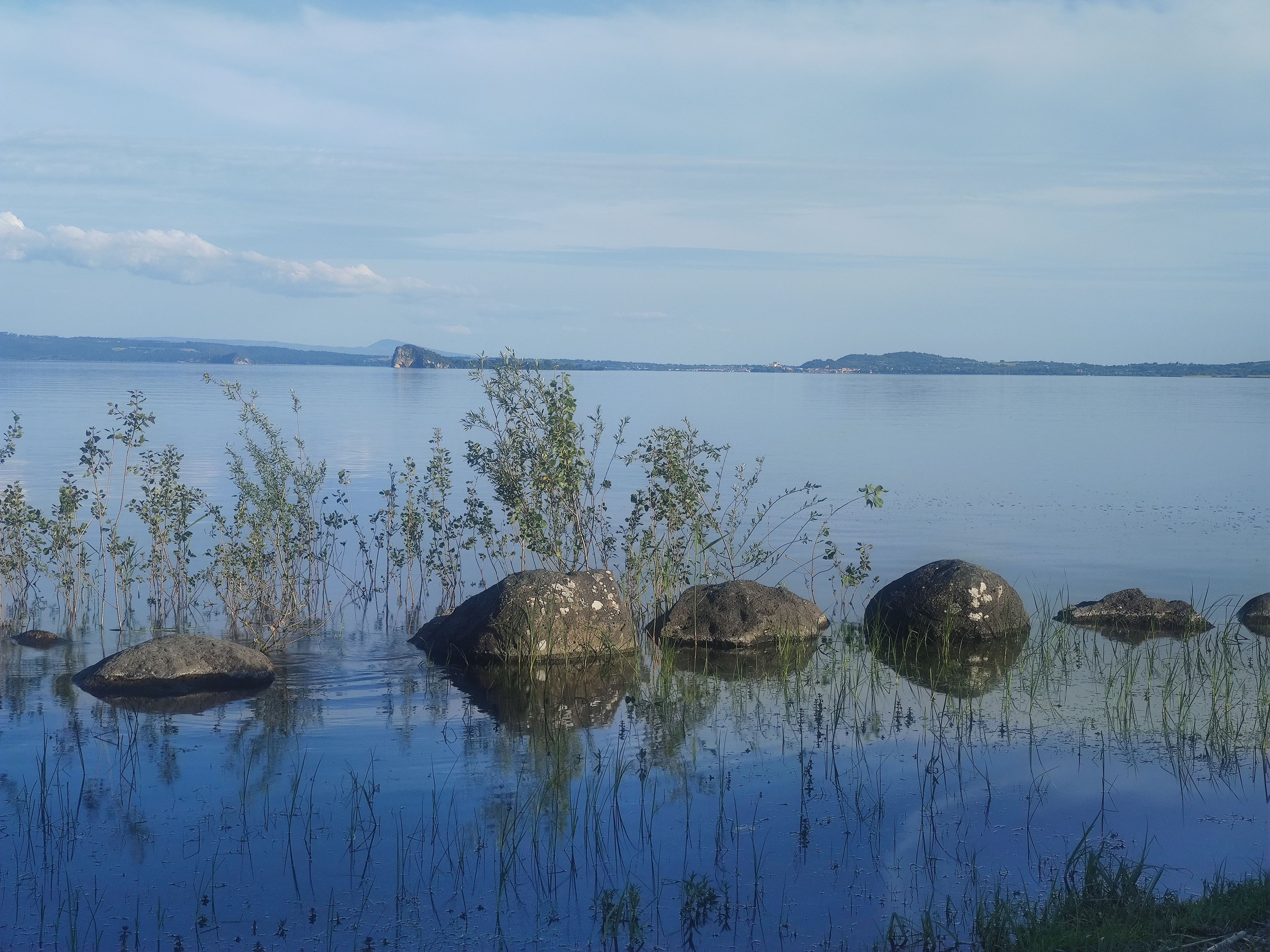 Un lac paisible, avec des rochers et de la végétation au premier plan, une rive lointaine parsemée de bâtiments et un ciel dégagé.