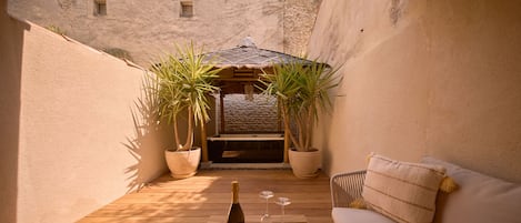 Una terraza de madera con un sofá, una mesa con una botella y vasos, y dos plantas en macetas.