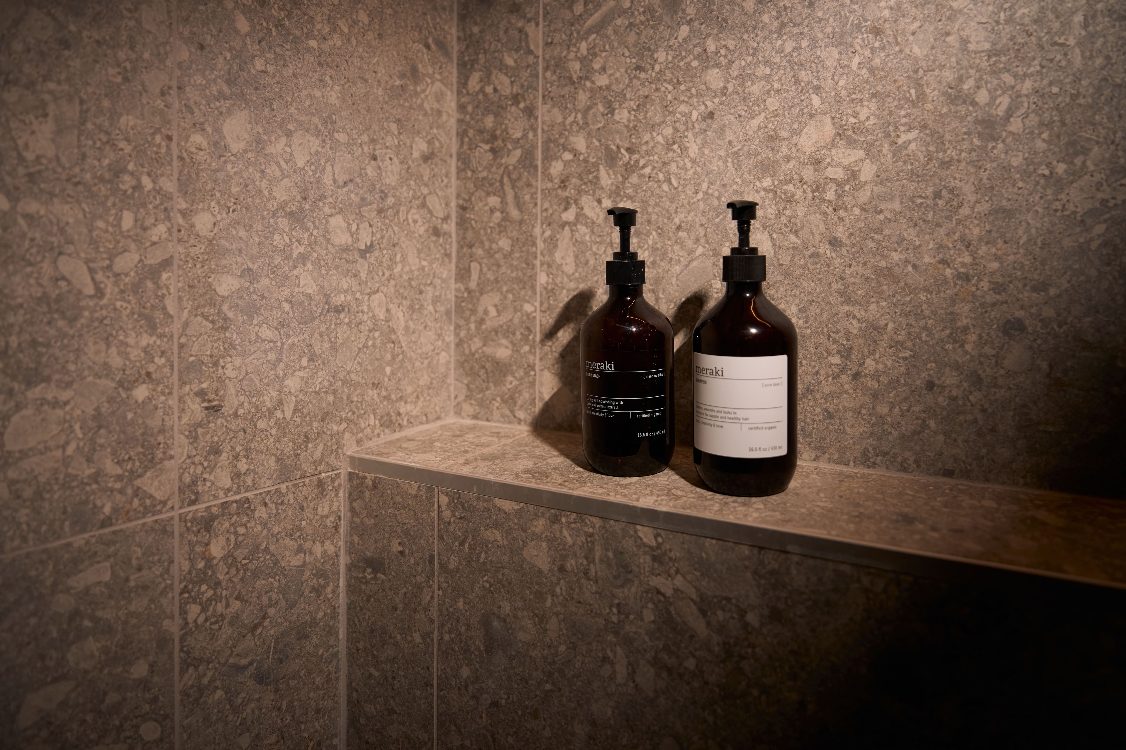 Deux bouteilles de shampoing et d’après-shampoing Meraki sont posées sur une étagère, devant un mur à la texture particulière.