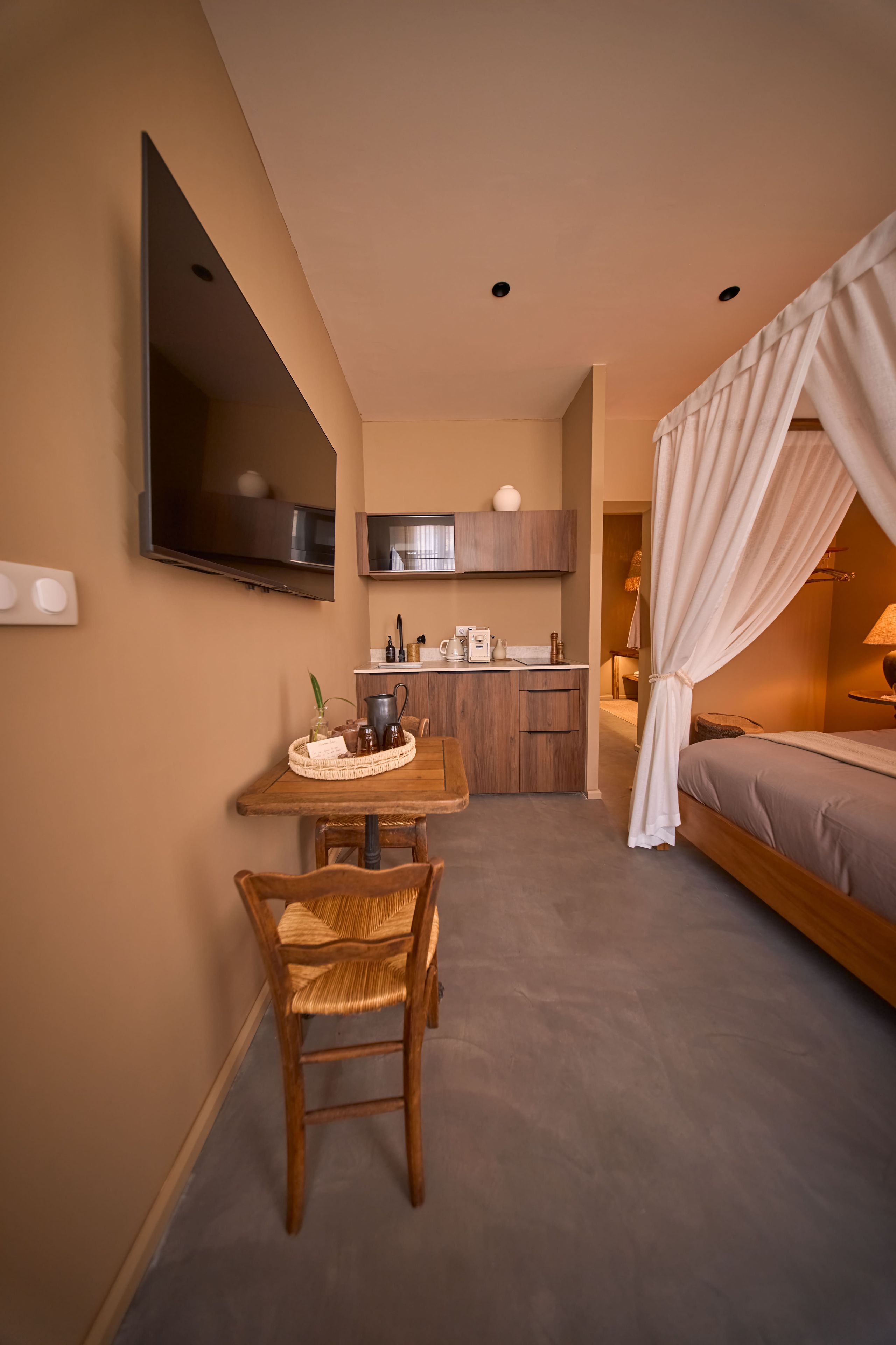 Een compacte hotelkamer met een bed, een kleine kitchenette, een eethoek met een tafel en stoelen, en een aan de muur bevestigde televisie.