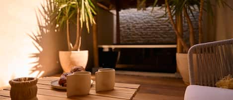 A wooden table with a plate of croissants and three ceramic cups, a woven basket, and a wicker chair in a cozy outdoor setting with potted plants and a stone wall.