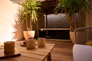 Una mesa de madera con un plato de croissants y tres tazas de cerámica, una cesta de mimbre y una silla de mimbre en un ambiente exterior acogedor con plantas en macetas y un muro de piedra.