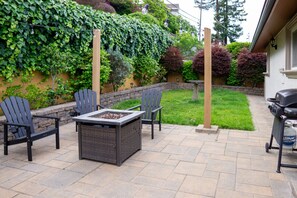 Eine Außenpatio mit einem Feuerkorbtisch, zwei Stühlen, einem Grill und einem Garten mit einer efeuüberrankten Mauer.