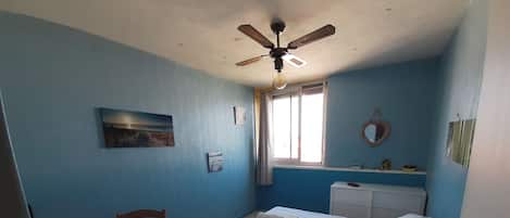 A room with a bed, a desk, a chair, a ceiling fan, a window, and a mirror.