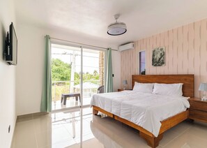 Une chambre à coucher comprenant un lit, une table de chevet, une lampe, un ventilateur de plafond, une fenêtre avec des rideaux et un appareil de climatisation.