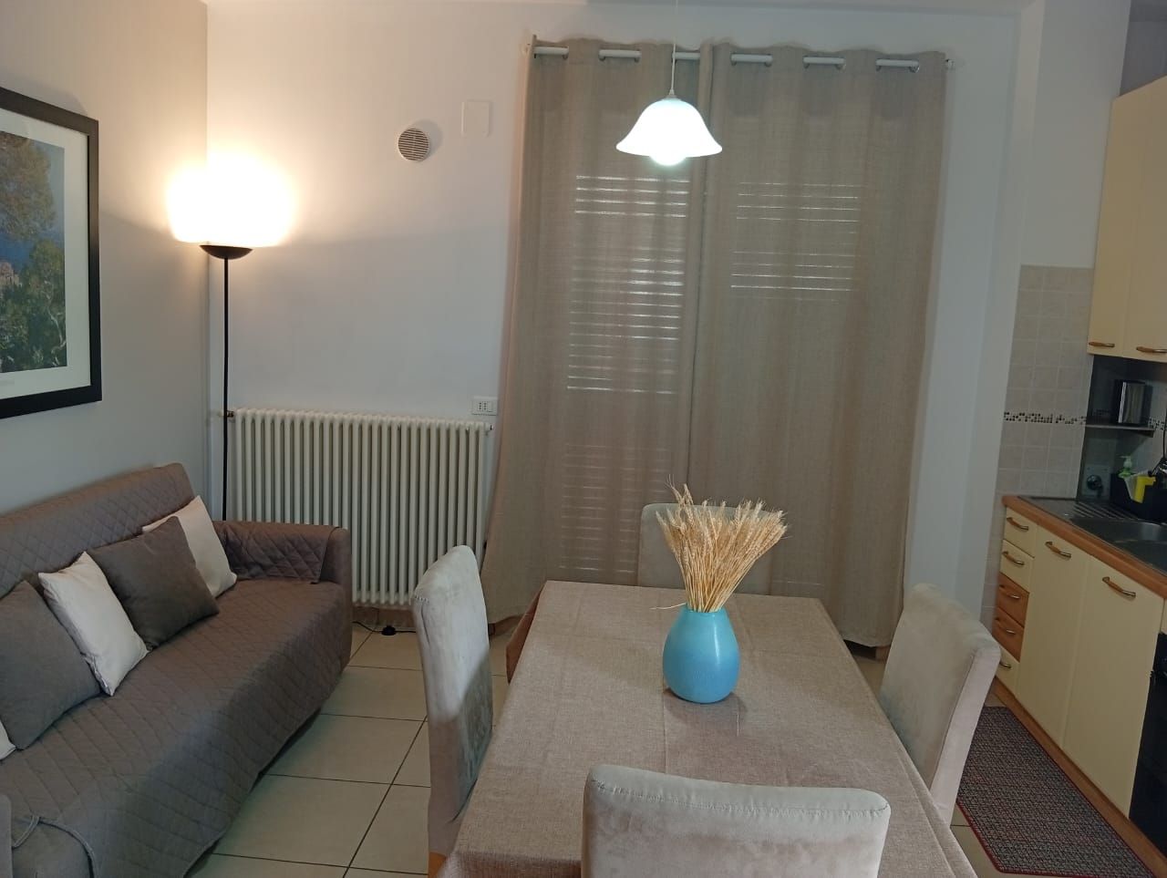Een woonkamer met een bank, een eettafel met stoelen, een blauwe vaas en een keuken met kasten.