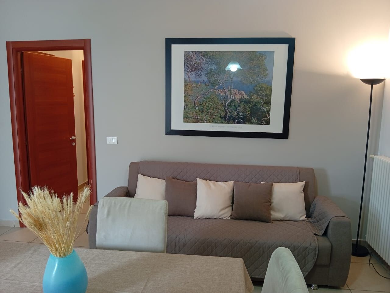 Un saló amb un sofà, un quadre al mur, una làmpada i un ram amb plantes seques.