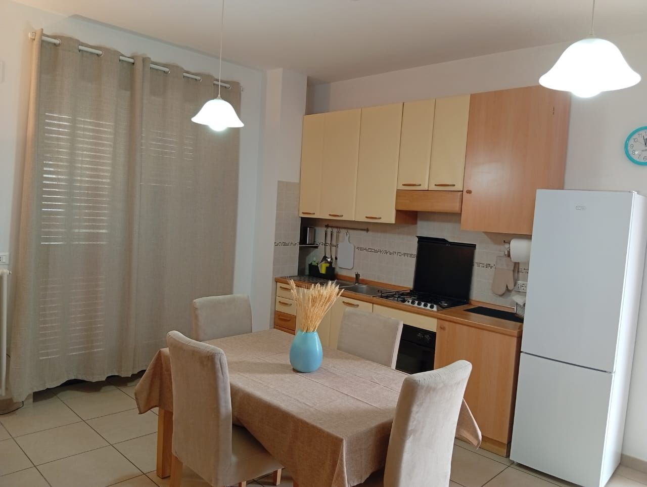 Angolo pranzo con tavolo apparecchiato per due, cucina con pensili in legno, fornello e frigorifero.