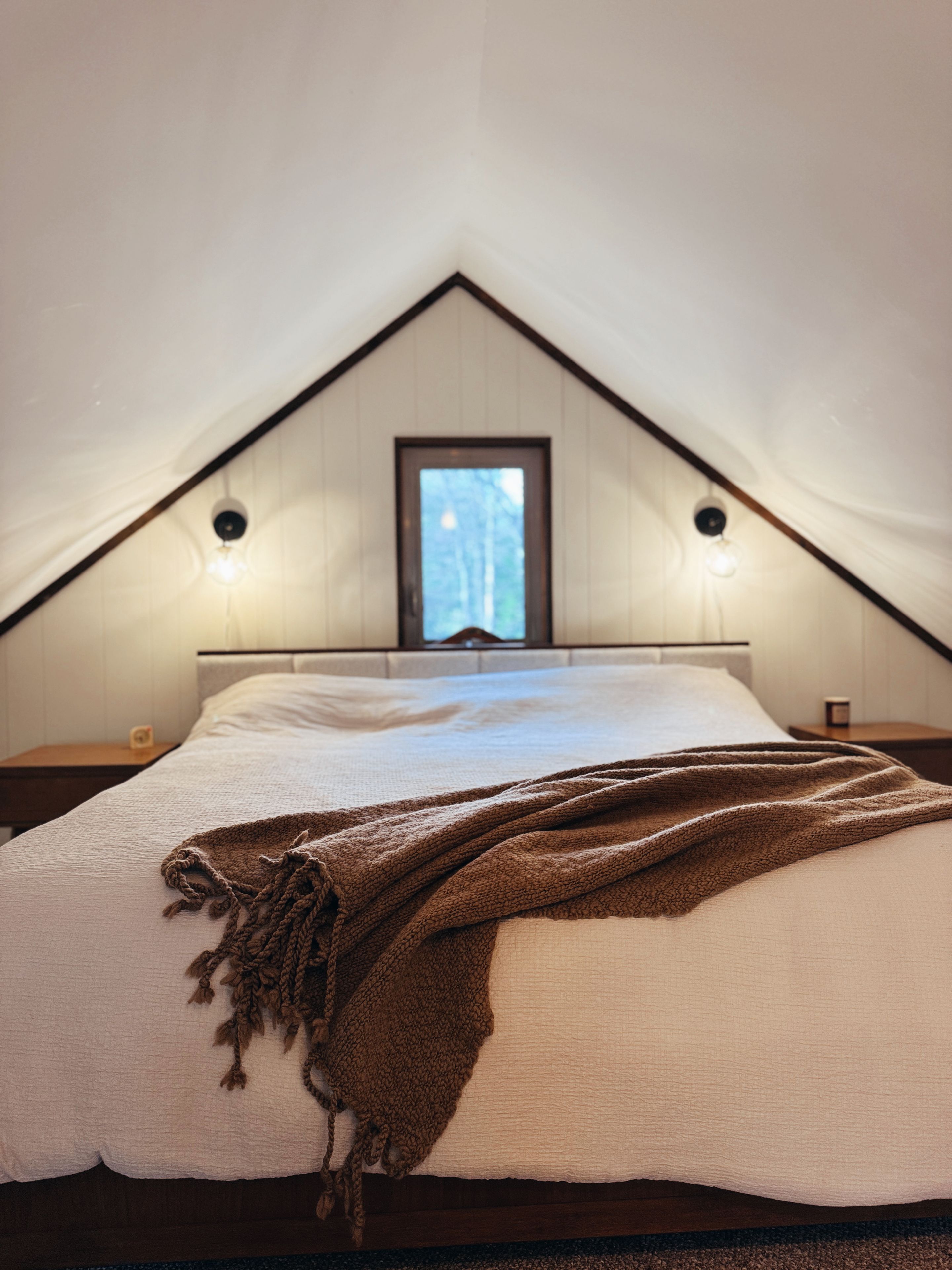 Une chambre avec un plafond en pente, un lit recouvert d’une couette blanche, un plaid marron et deux lampes fixées au mur.