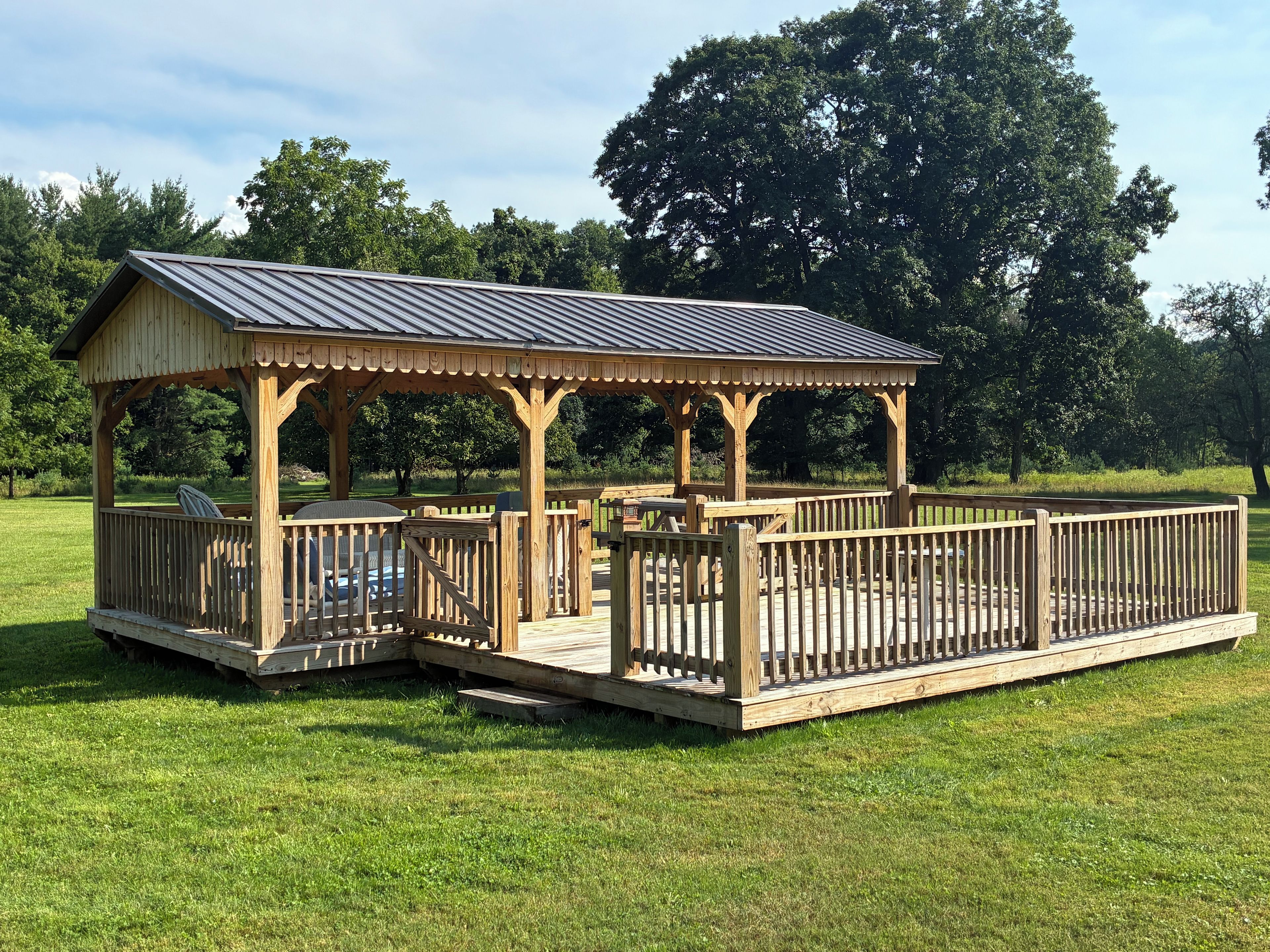 A wooden gazebo with a metal roof, surrounded by a wooden deck and a grassy area.