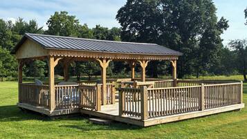 A wooden gazebo with a metal roof, surrounded by a wooden deck and a grassy area.