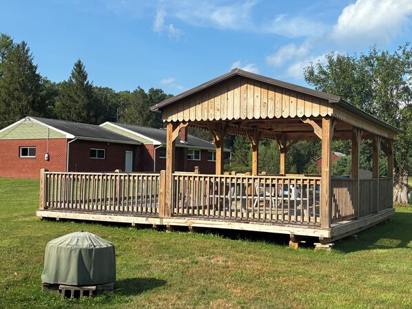 A wooden gazebo with a deck, surrounded by a grassy field and a brick building in the background.