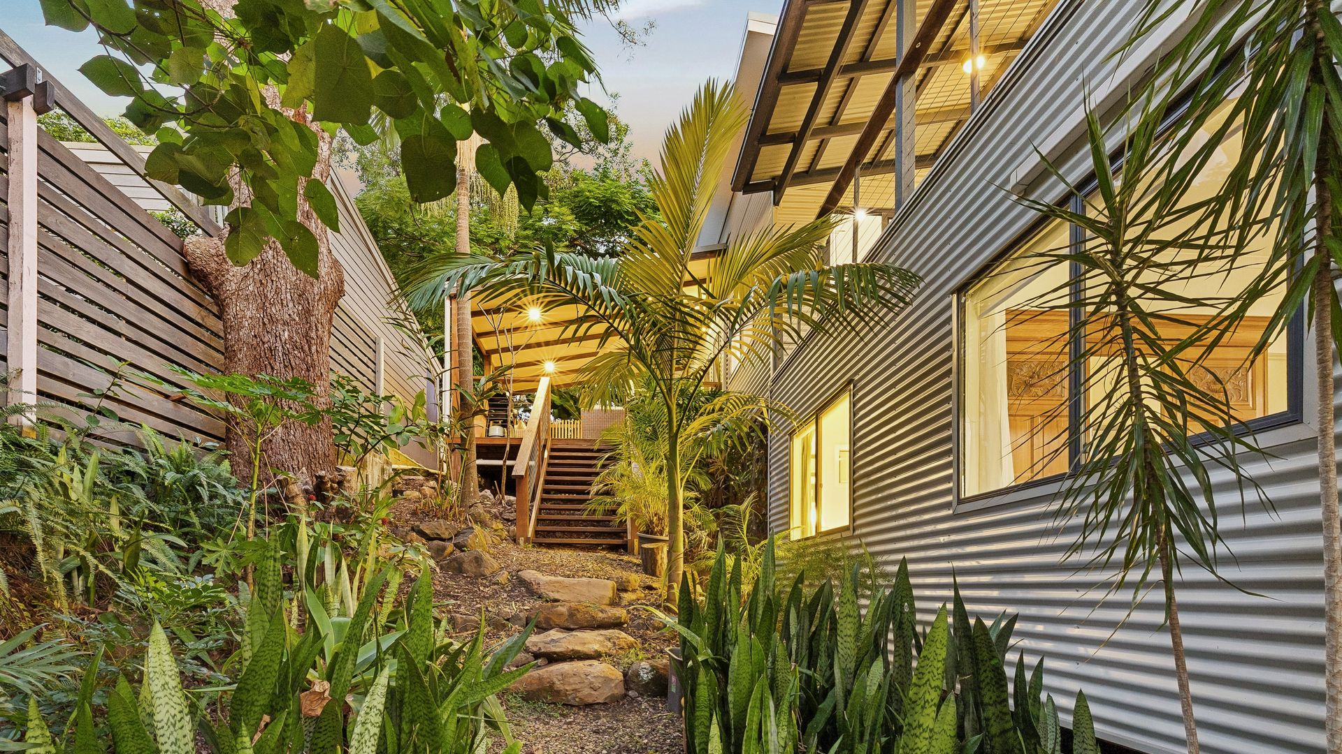 Een modern gebouw met een houten terras, een trap naar een overdekt gedeelte en verschillende tropische planten.