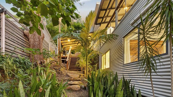 Un edificio moderno con una terrazza in legno, una scala che conduce a un'area coperta e diverse piante tropicali.