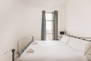 Ein ordentlich gemachtes Bett mit weißen Bettwäsche und einem Metallgestell, eine Nachttischlampe und ein Fenster mit Vorhängen.