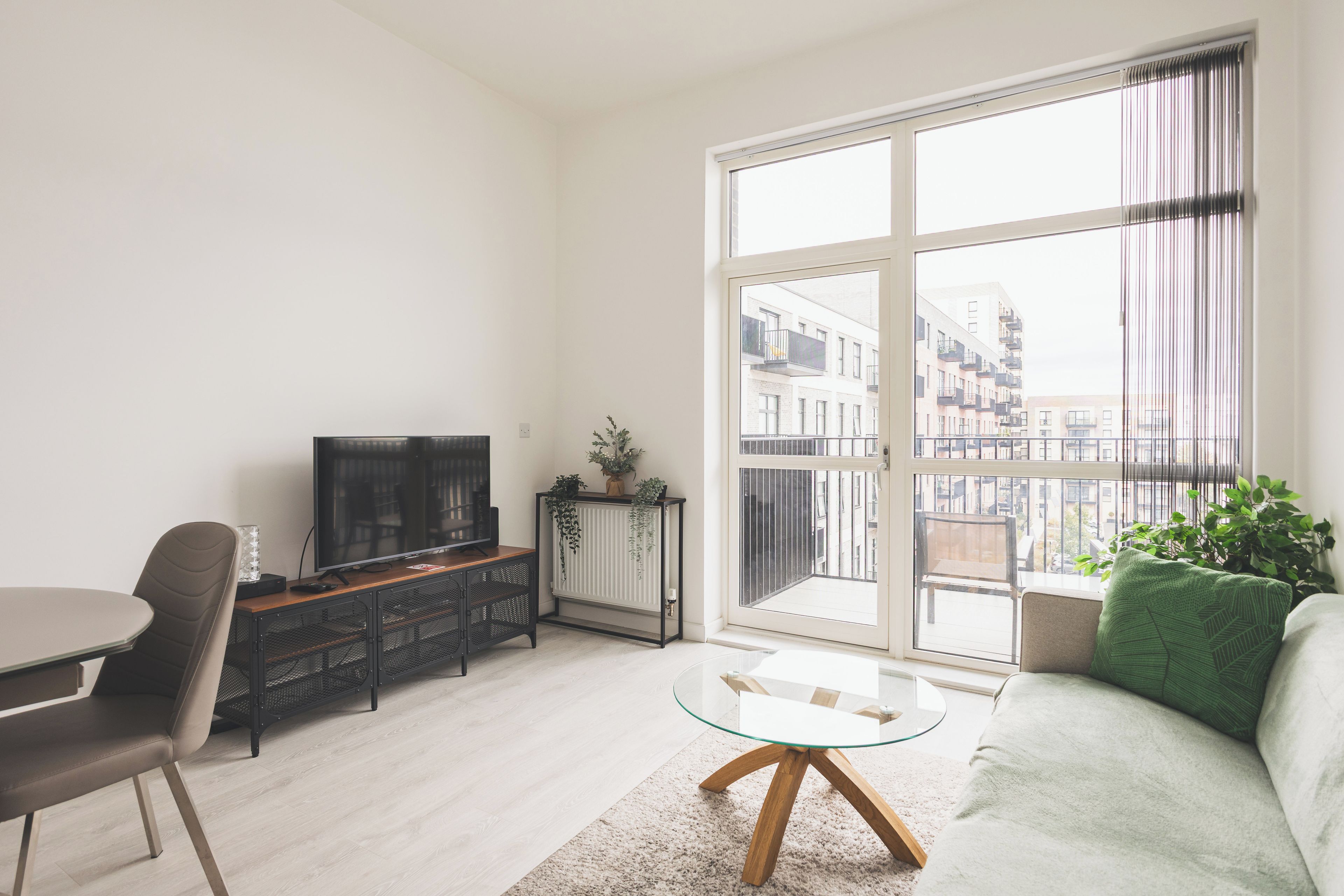 Una sala de estar moderna con televisor, asientos, una mesa de centro de vidrio y un ventanal con vista a edificios.