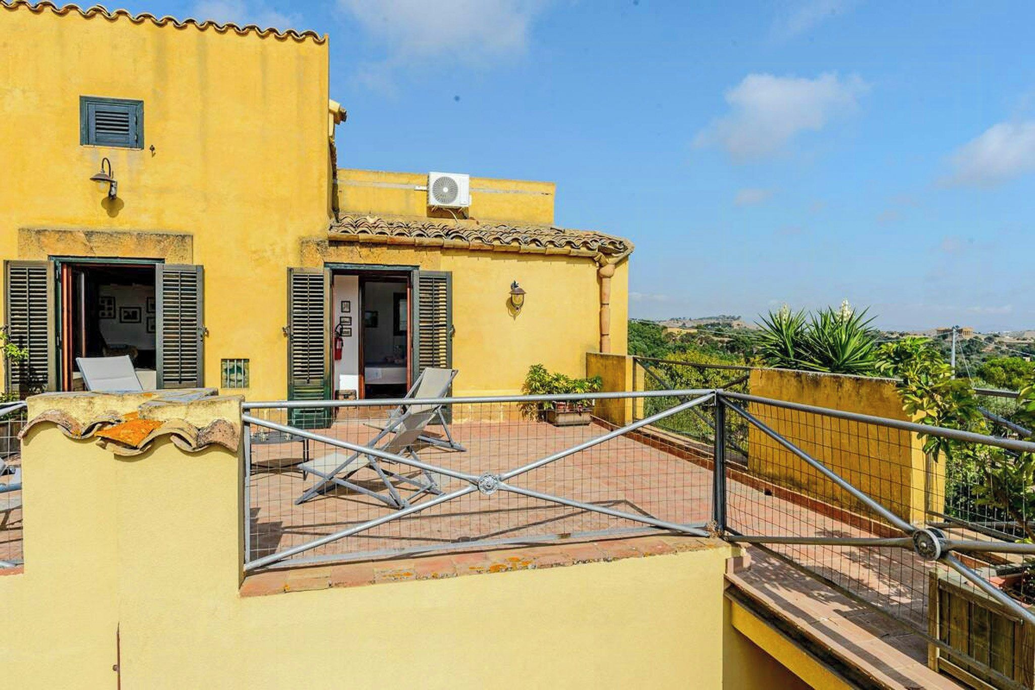 A yellow building with a terrace, a metal railing, and a view of the landscape.