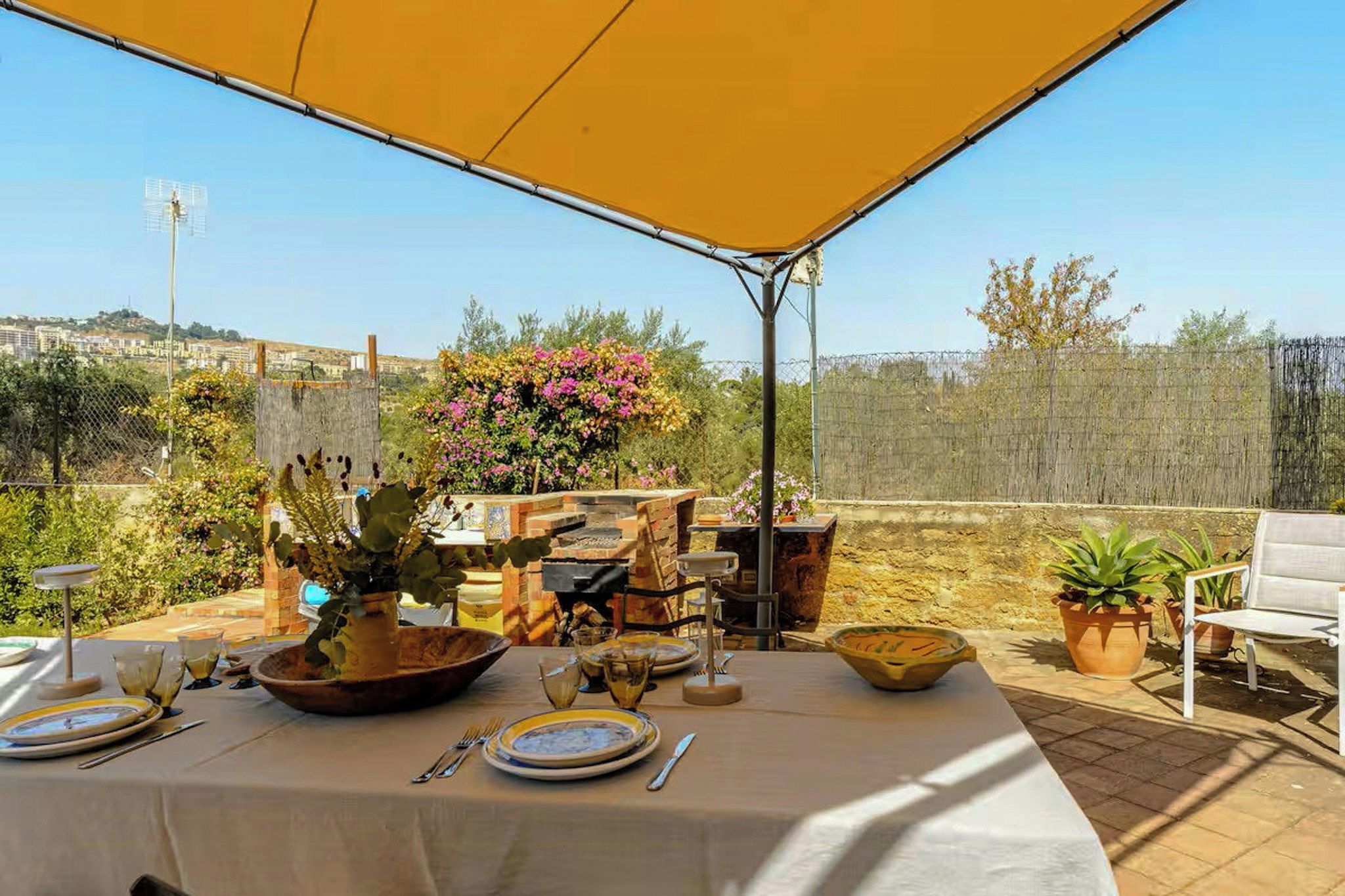 Un área de comedor al aire libre con una mesa para dos, rodeada de macetas con plantas y un horno de ladrillo.