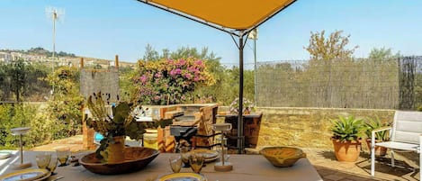Un área de comedor al aire libre con una mesa para dos, rodeada de macetas con plantas y un horno de ladrillo.