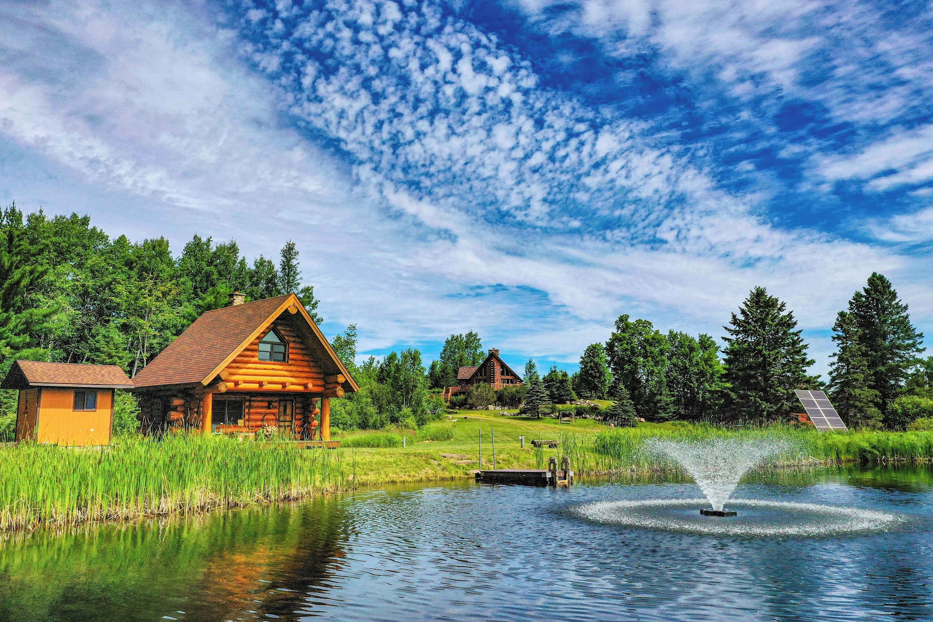 A log cabin with a pond and a fountain, surrounded by trees and a clear sky.