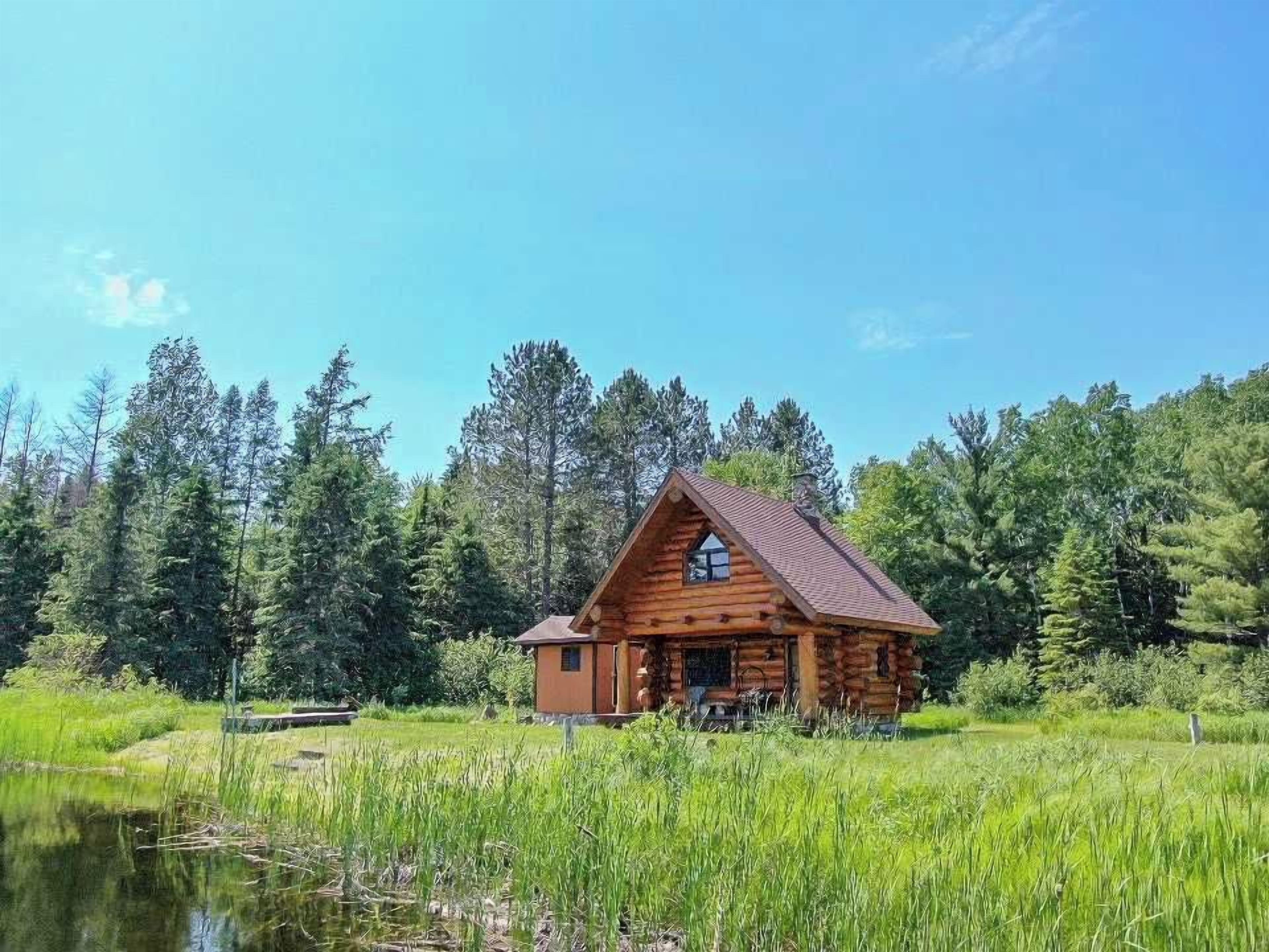 A wooden cabin with a sloped roof, situated by a body of water surrounded by dense forest.
