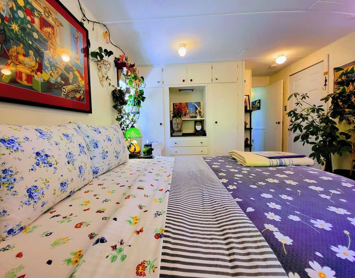 Et soveværelse med en sofa, en seng med blomstret og stribet sengetøj, et natbord med en lampe og en vægmonteret hylde med dekorative genstande.