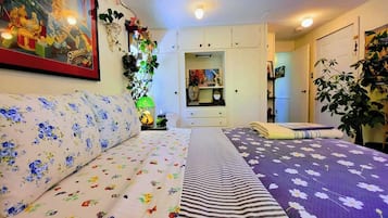 Et soveværelse med en sofa, en seng med blomstret og stribet sengetøj, et natbord med en lampe og en vægmonteret hylde med dekorative genstande.