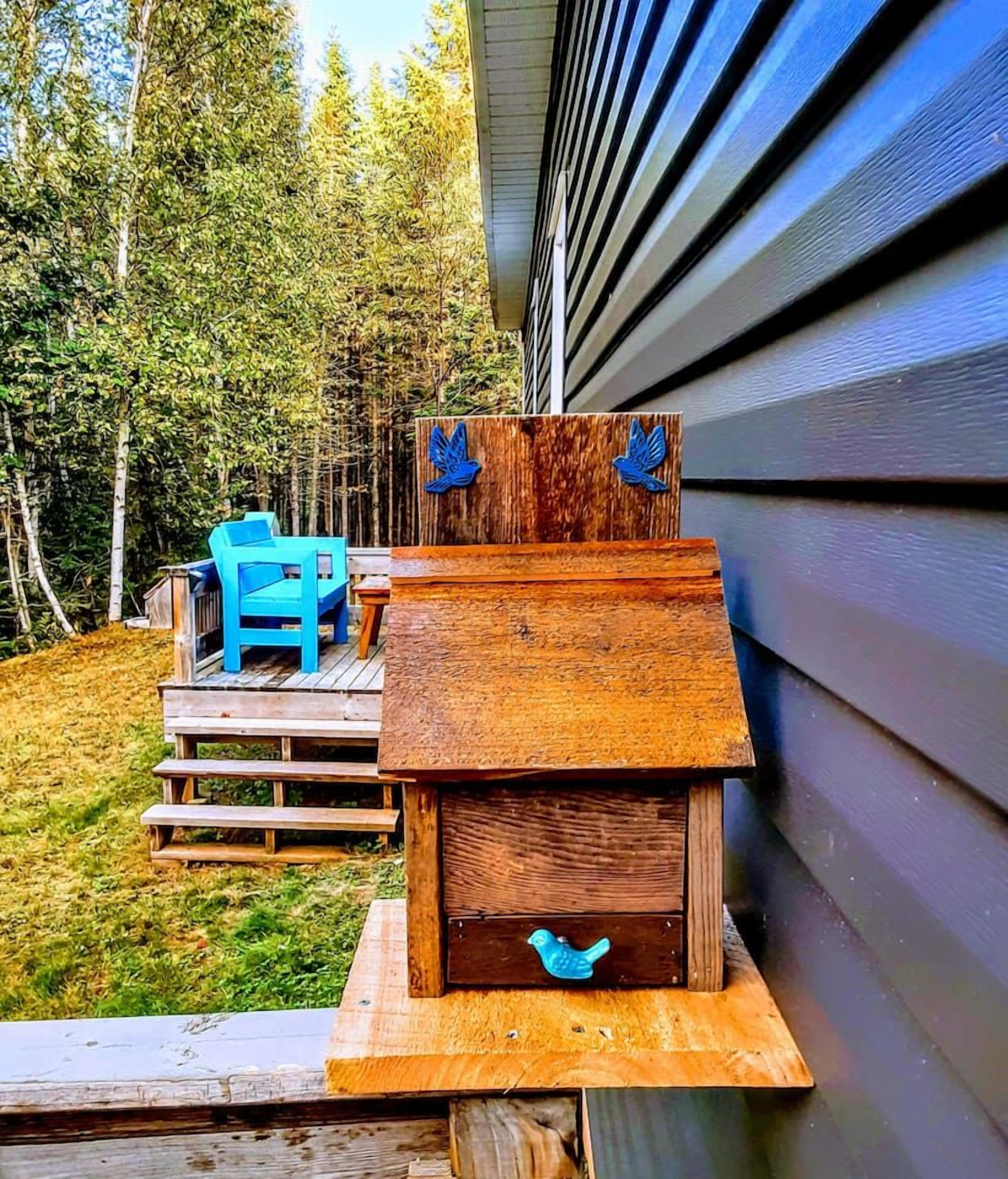A wooden birdhouse attached to a house with a deck and a blue chair.