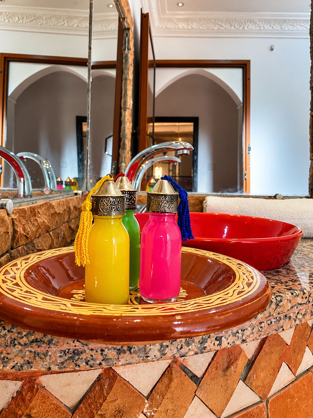 A bathroom with a marble sink, a red basin, and three colorful bottles with tassels.