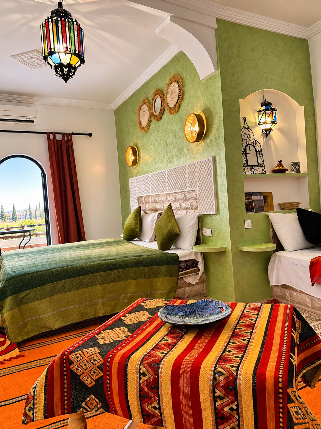 Ein Hotelzimmer mit zwei Betten, einer grünen Akzentwand, dekorativen Wandleuchten, einem gemusterten Teppich und Blick durch ein Fenster.