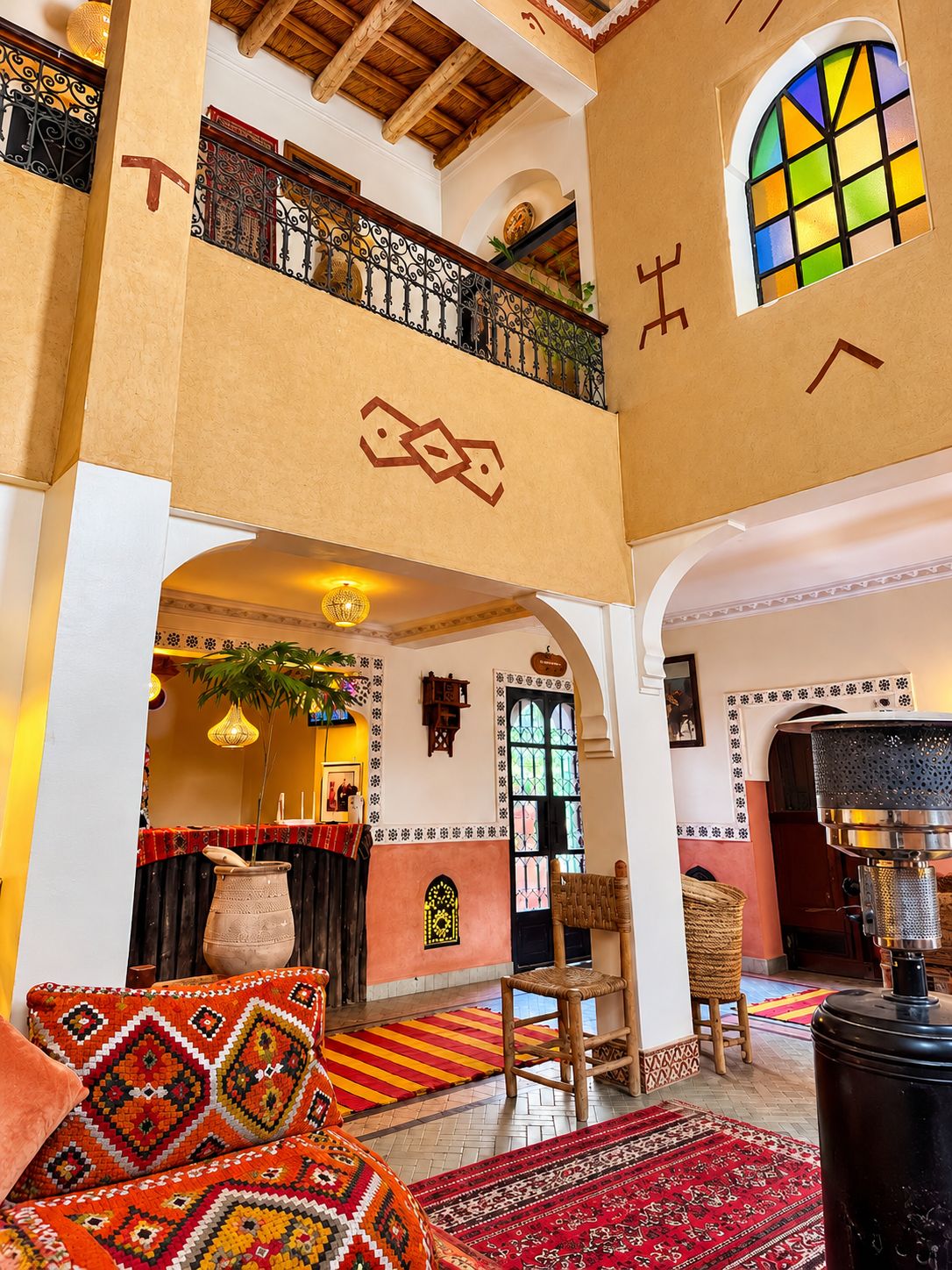 A hotel lobby with a high ceiling, a balcony with ornate railings, a large window with colorful stained glass, and a traditional woven chair.