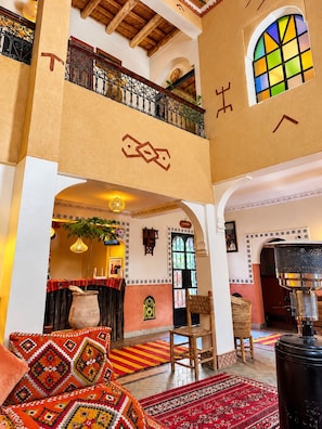 A hotel lobby with a high ceiling, a balcony with ornate railings, a large window with colorful stained glass, and a traditional woven chair. - auberge tissadrine (Ait Sedrate Jbel El Soufla)