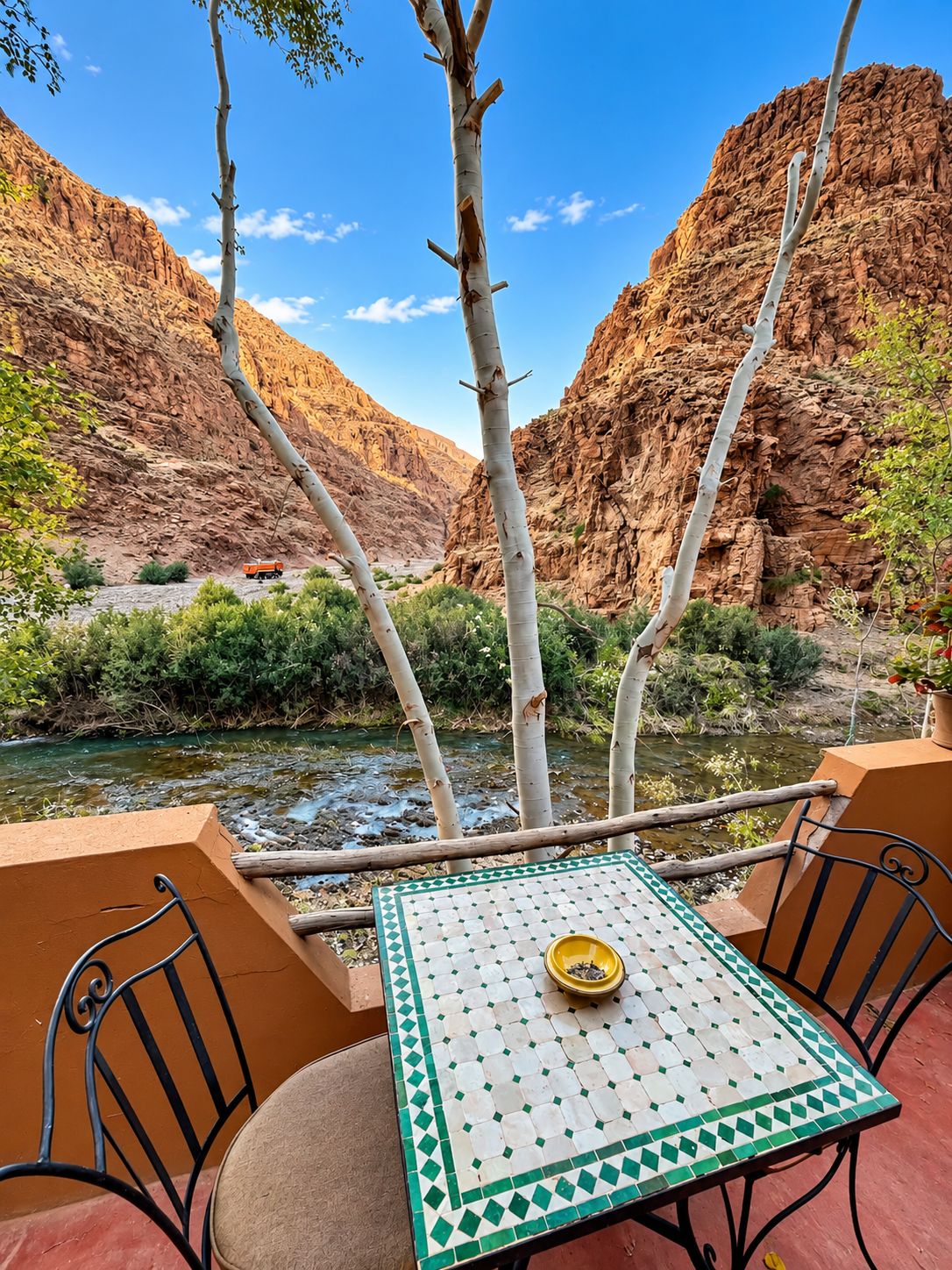 A table with a patterned tablecloth, a bowl, and chairs, set against a backdrop of a canyon with a river.