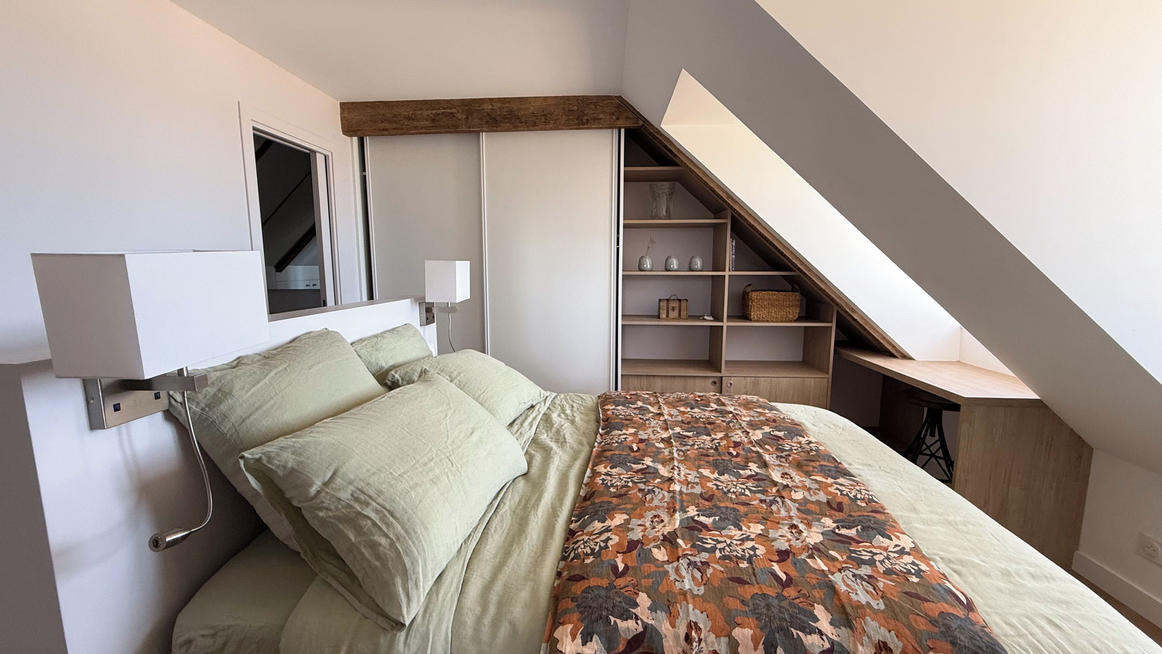 A bedroom with a bed, a bedside table, a lamp, a window, and a built-in shelving unit.