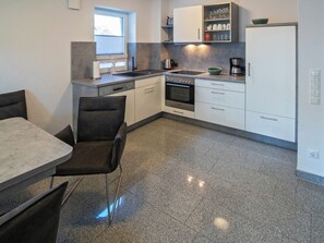 A modern kitchen with a dining area, featuring a built-in oven, a coffee maker, and a window with blinds. - Holiday Home With 1 Bedroom (Esens)