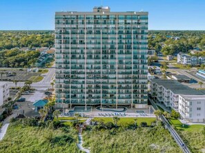 A multi-story hotel with a modern glass facade, a central courtyard with a pool, and a parking area. - Oceanfront 3BR- Windy Hill Dunes 705- Lazy River (North Myrtle Beach)