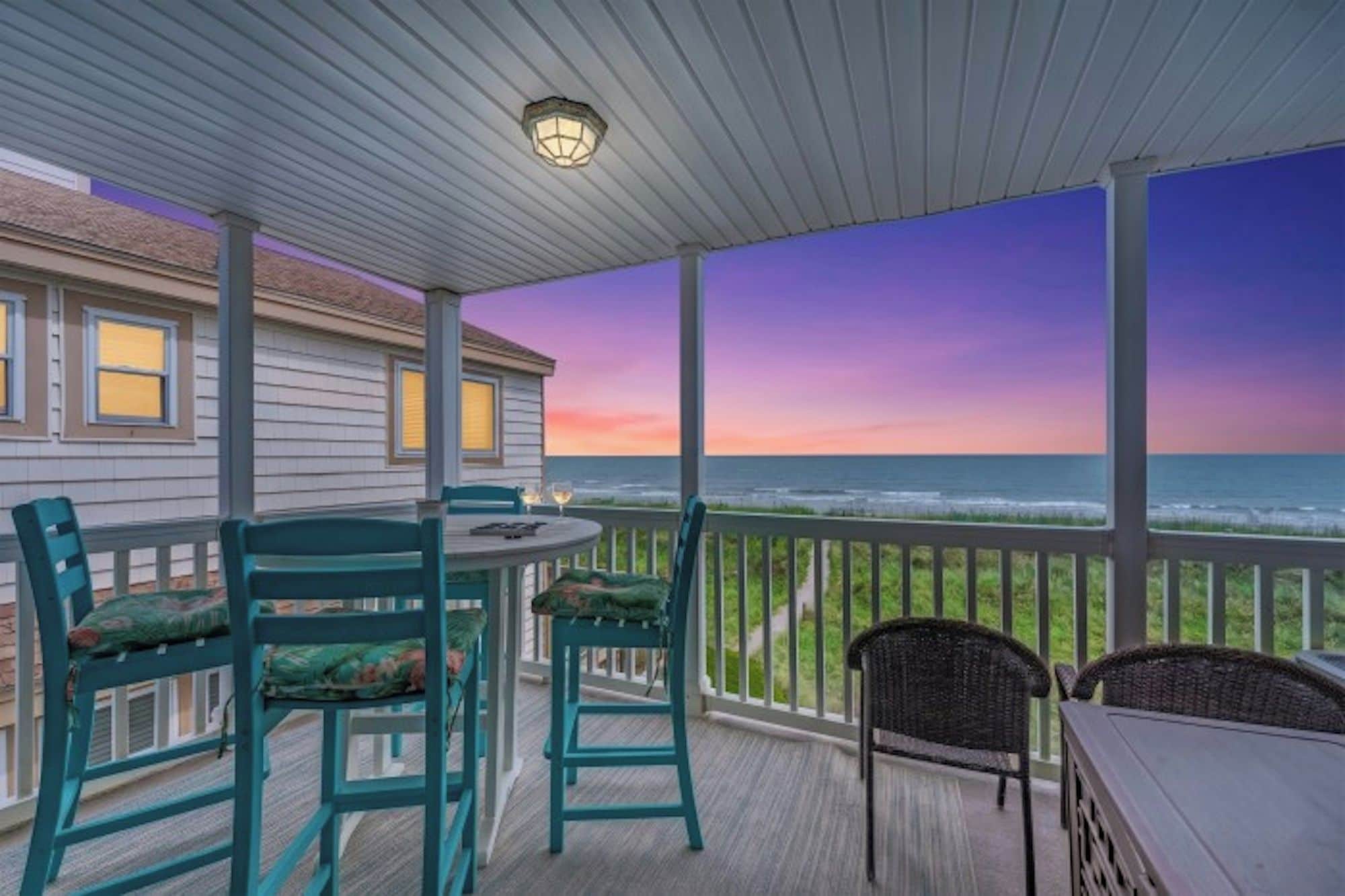 A covered outdoor area with a table and chairs, overlooking a beach with a sunset view.