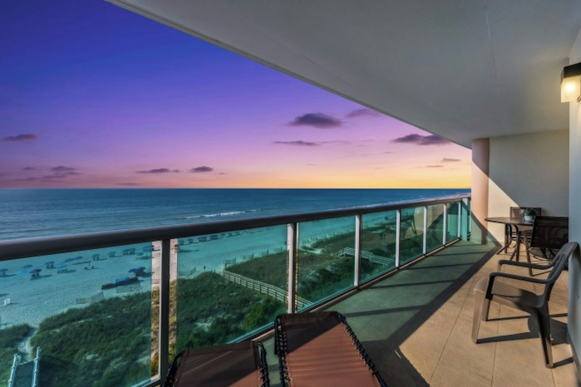 A balcony with a view of the ocean at sunset, a table with chairs, and a railing.