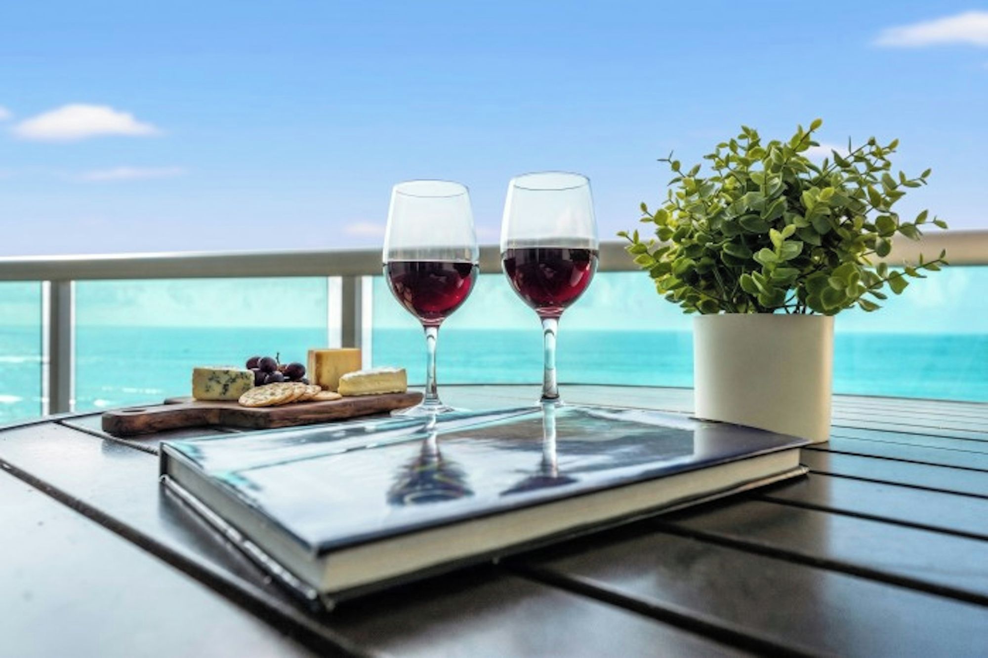 Una mesa con un libro, dos copas de vino tinto, una planta pequeña y una tabla de quesos con galletas y uvas.