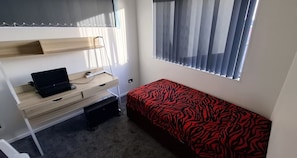 Ein kleines Zimmer mit einem Bett, einem Schreibtisch mit Laptop und einem Fenster mit Jalousien.