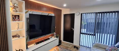 A modern living room with a large flat-screen TV, a sofa, a coffee table, and a window with blinds.