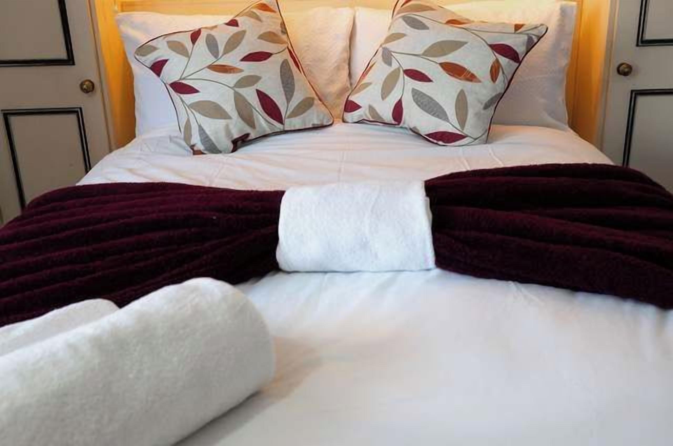 A neatly made bed with a white comforter, two pillows with a leaf pattern, and a dark red blanket.