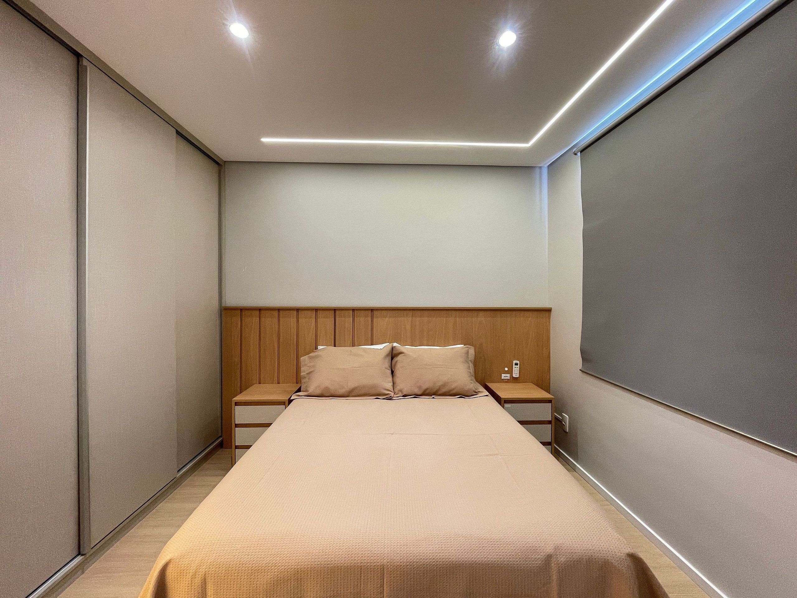 A narrow hotel room with a single bed, a wooden headboard, a bedside table with a phone, and a wall-mounted light fixture.