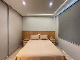 Quarto de hotel estreito com uma cama de solteiro, cabeceira de madeira, mesa de cabeceira com telefone e luminária fixada na parede.