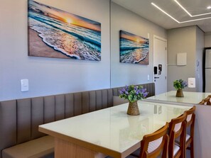 Un comedor moderno con una mesa de vidrio, sillas de madera y un banco. Hay dos cuadros colgados en la pared que representan una escena de playa.